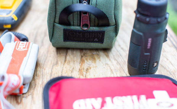 A Boreal Grab Bag showing the TOM BIHN Design Lab label on a picnic table with a headlamp, First Aid Kit, and flashlight.